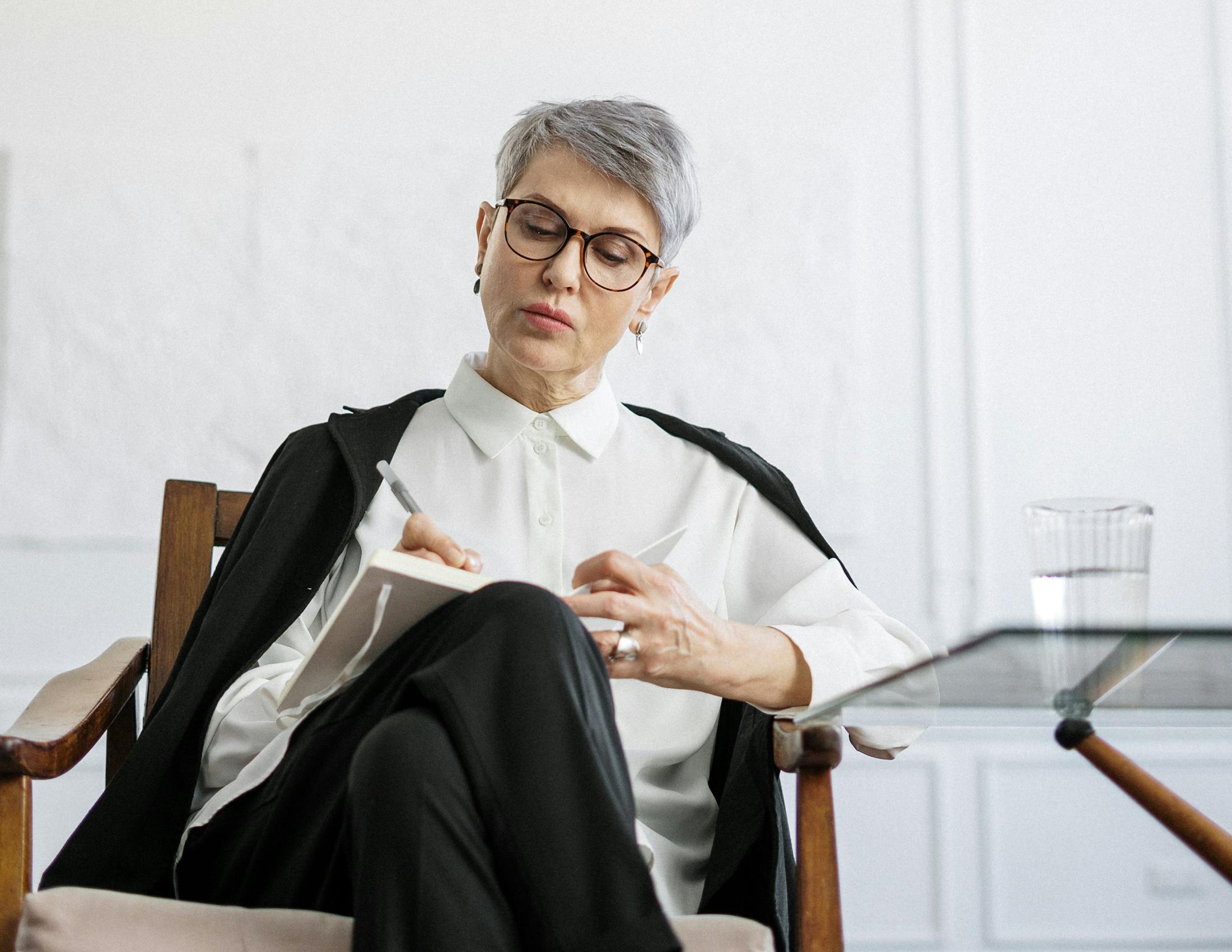 A grey haired therapist with glasses taking notes while sitting on an armchair with a white background.