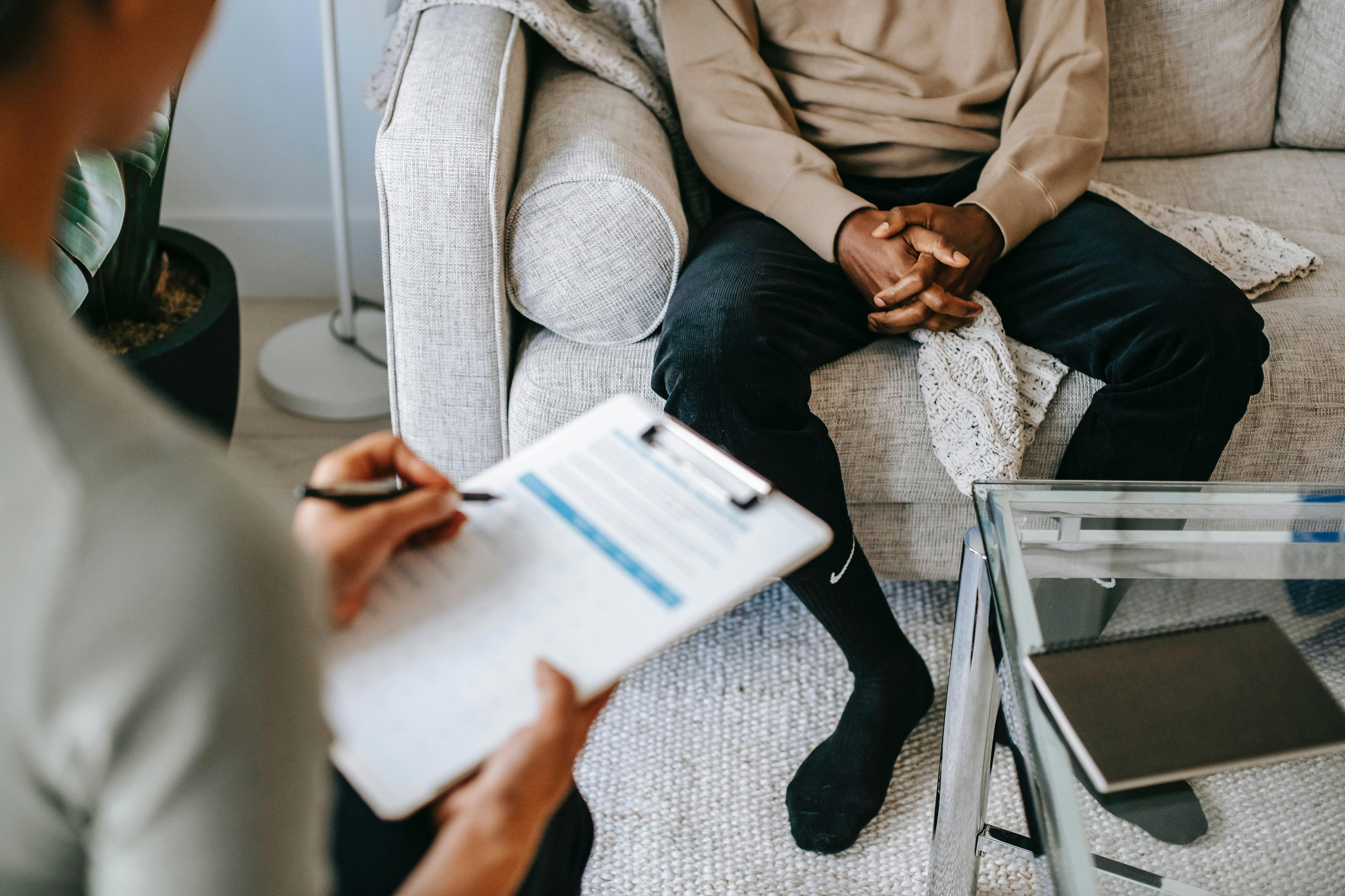 Therapist writing notes while sitting down in front of a patient who is sitting on a sofa.